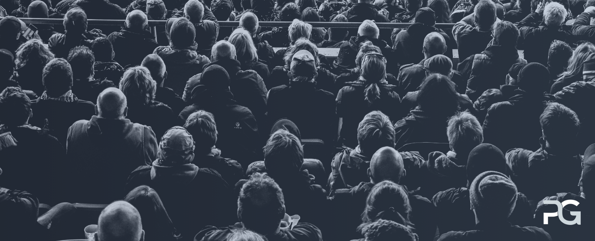 A large crowd of people seated closely together, viewed from behind, attending an event or presentation in a dimly lit venue.